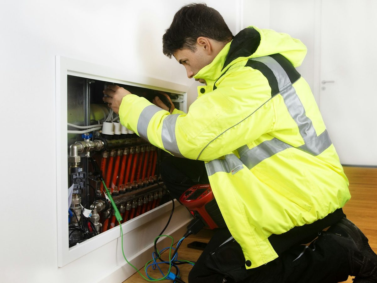 An engineer in a hi-vis jacket working on a heating manifold set into a wall.
